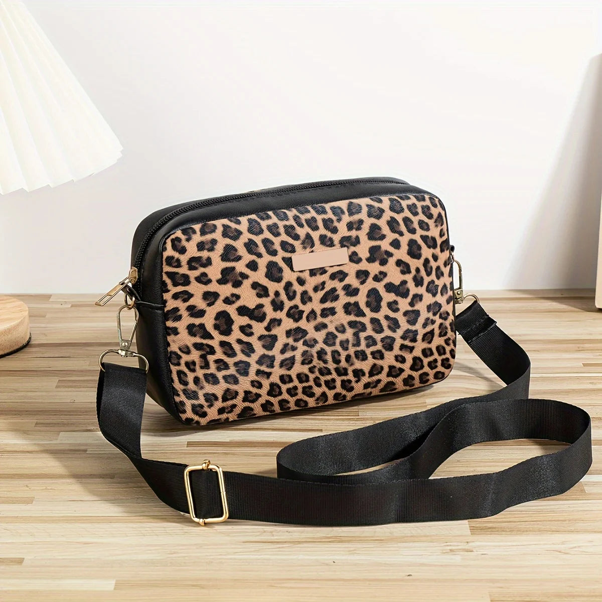 A black leopard print crossbody bag with a gold buckle is displayed on a wooden surface.
