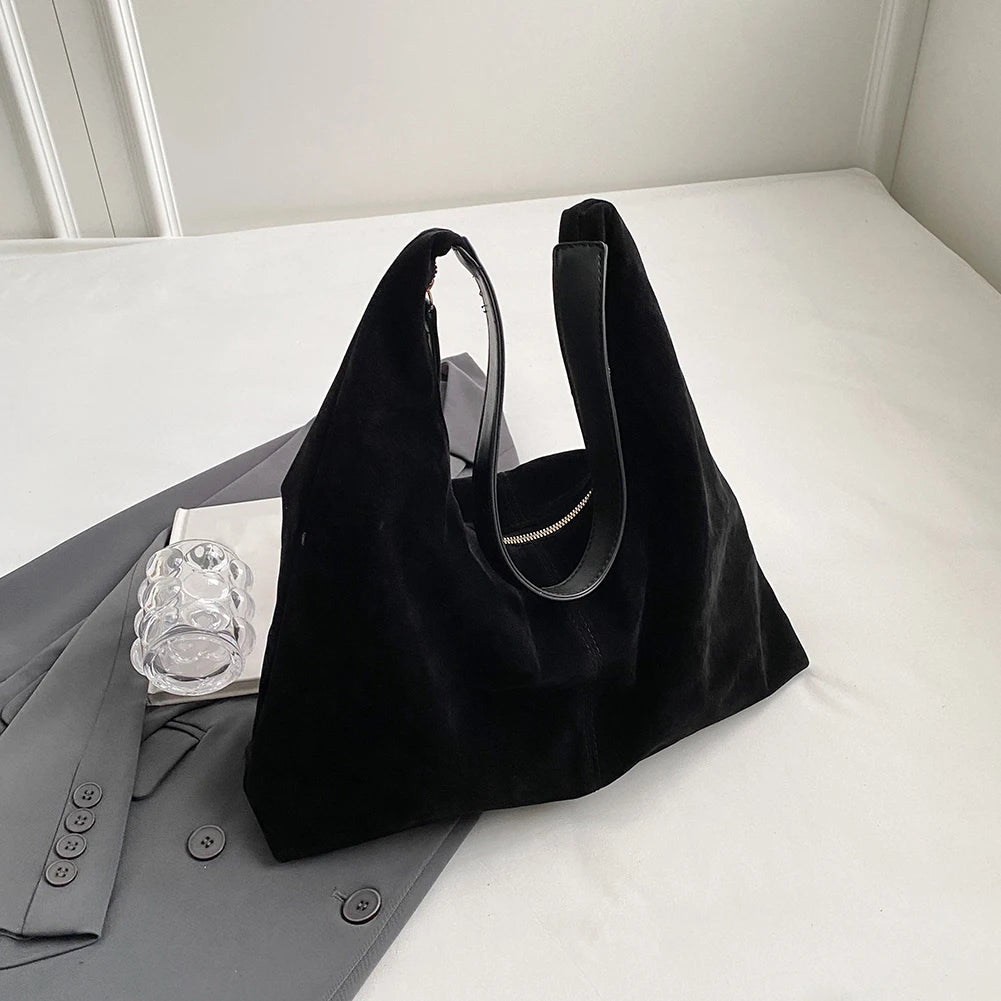 A black suede slouchy shoulder bag resting on a white surface next to a gray blazer.