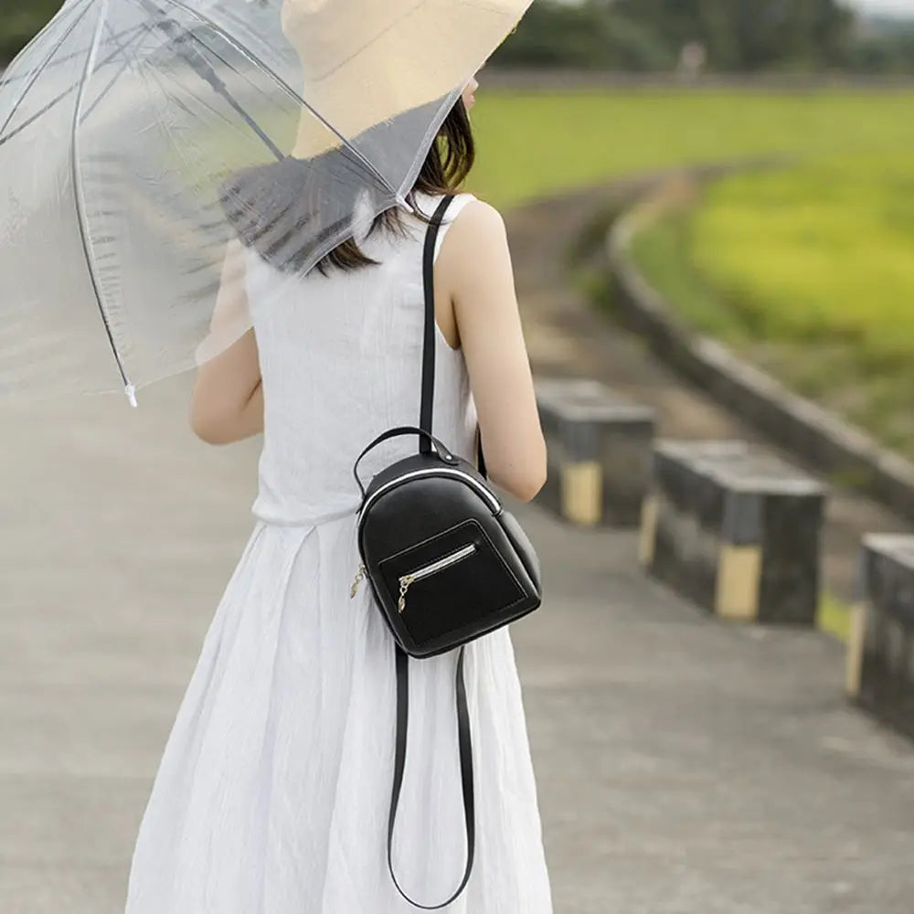 A woman in a white dress holds a clear umbrella and carries a black waterproof mini backpack while walking on a path.