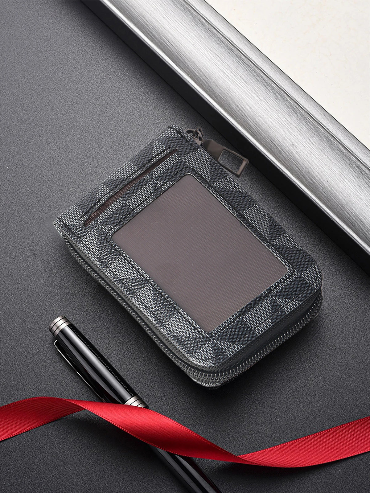 Schwarzes Kunstleder-Kartenträger mit Reißverschluss auf dunkler Oberfläche neben einem Stift und einem silbernen Rahmen.