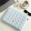 A light blue quilted laptop sleeve is shown next to a silver laptop, a perfume bottle, and a white cup on a white surface.