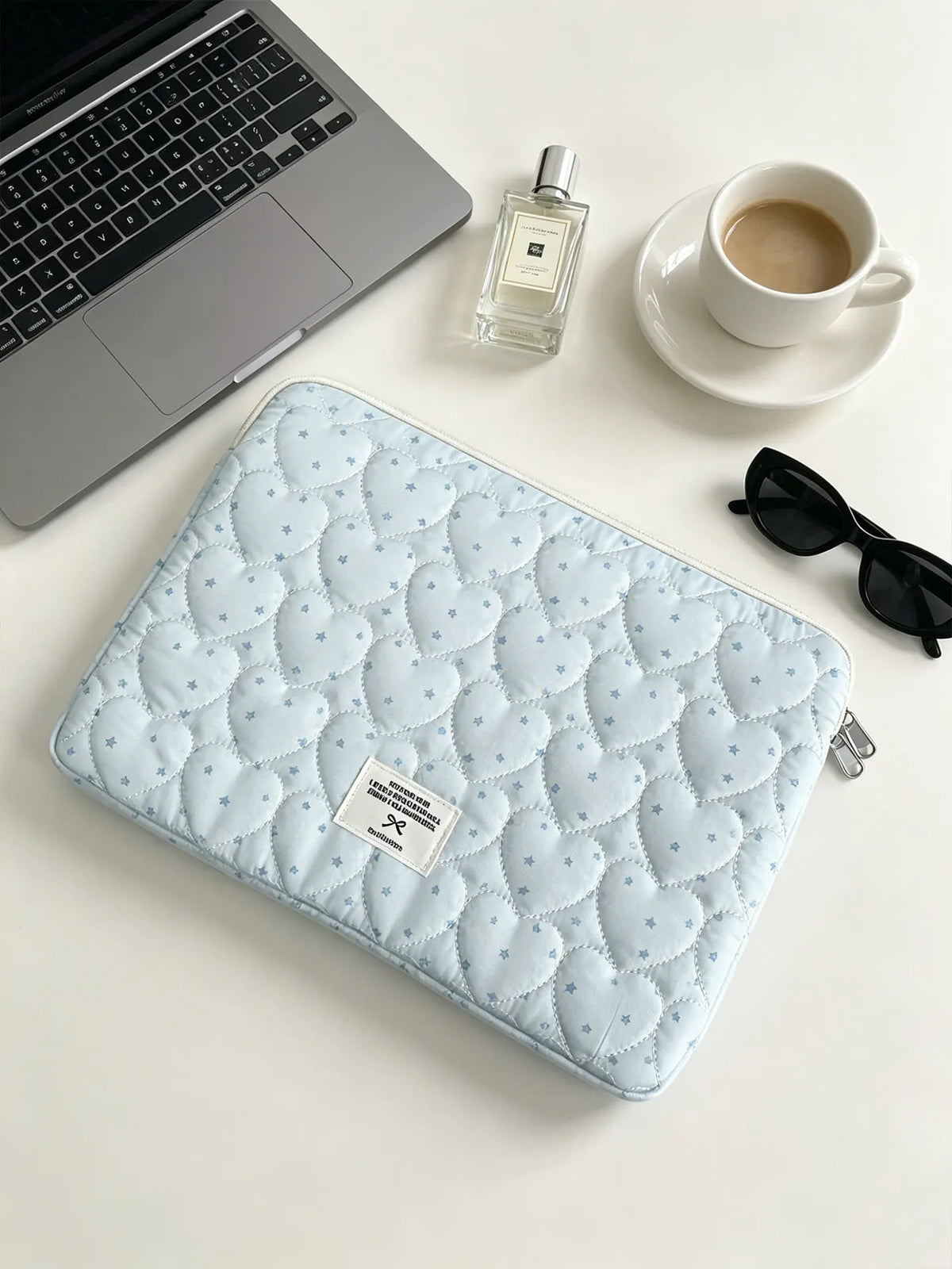 A light blue quilted laptop sleeve is shown next to a silver laptop, a perfume bottle, and a white cup on a white surface.
