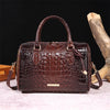 A dark brown crocodile pattern leather crossbody tote bag with a handle resting on a light surface next to a piece of driftwo