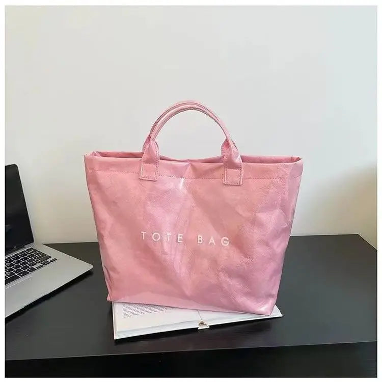 A pink satin tote bag is displayed on a dark surface next to a laptop and some papers.
