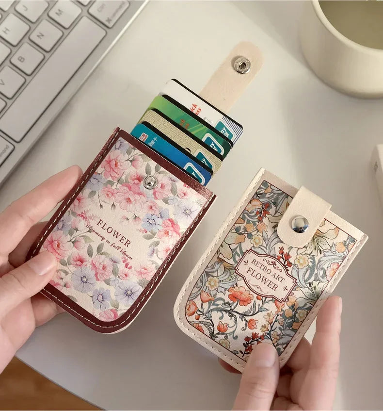 A hand holds a floral patterned leather card wallet next to a stack of credit cards on a white desk.