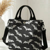 Black tote bag with a white bat print and black strap resting on a white surface.