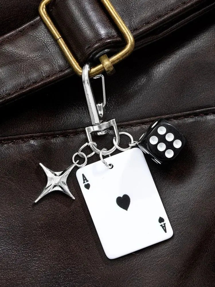 Llavero de plata con una carta blanca y un dado de póker negro, sujeto a una correa de cuero marrón.