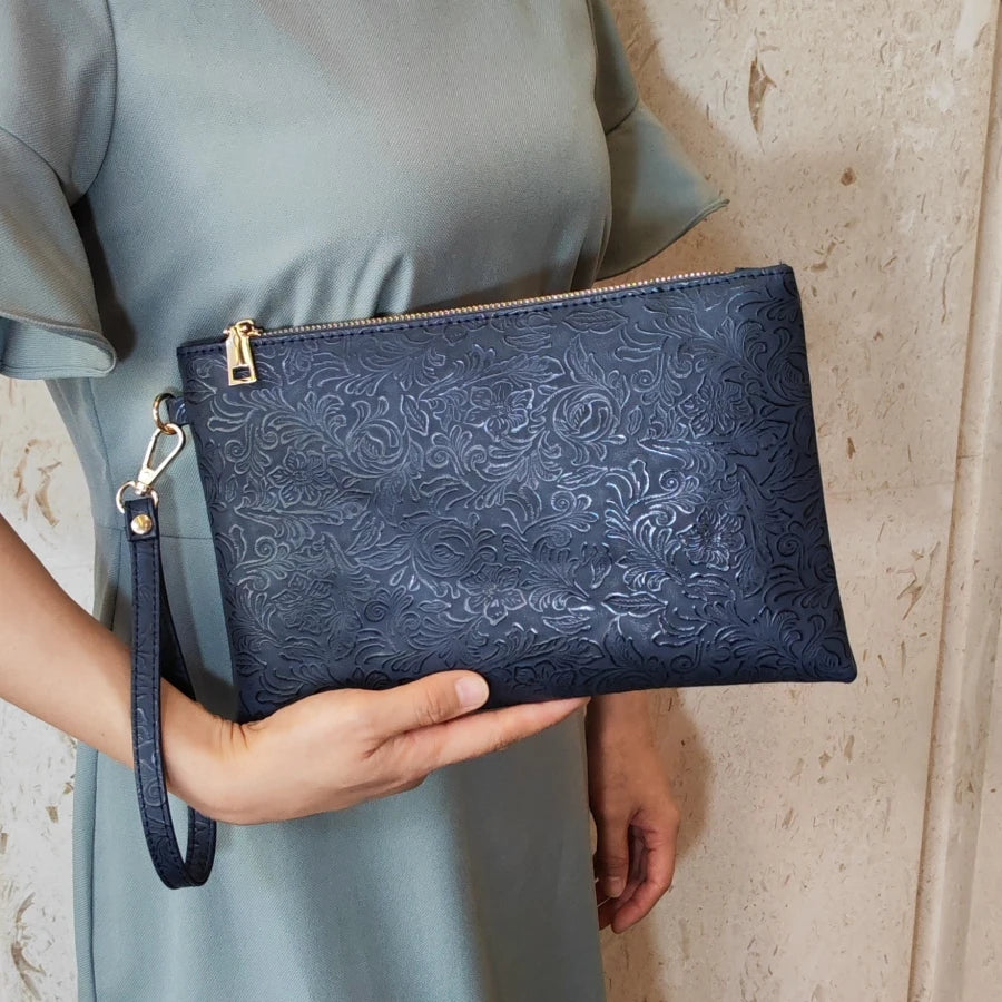 Una mano sostiene un bolso de cuero sintético azul oscuro con estampado floral contra un vestido verde claro.