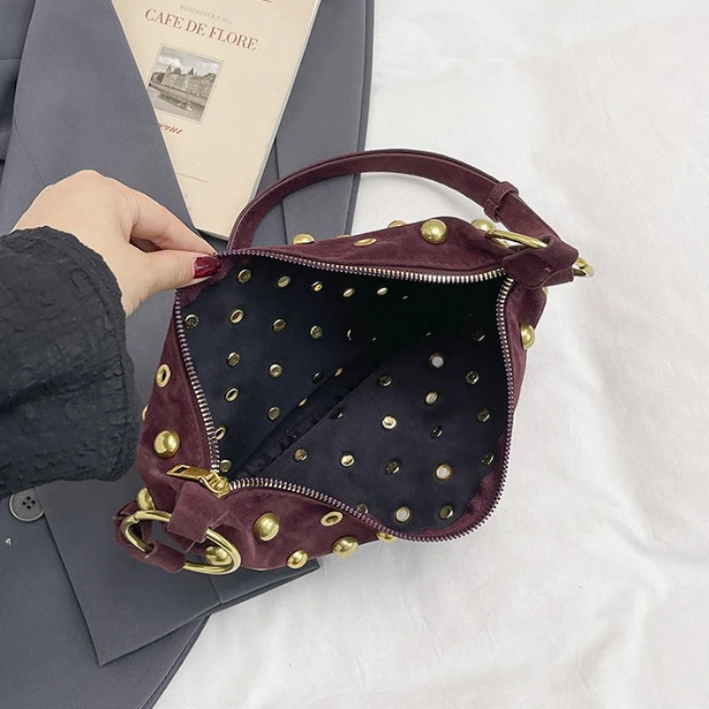 A dark burgundy faux suede hobo crossbody bag with gold rivet accents resting on a gray folder.