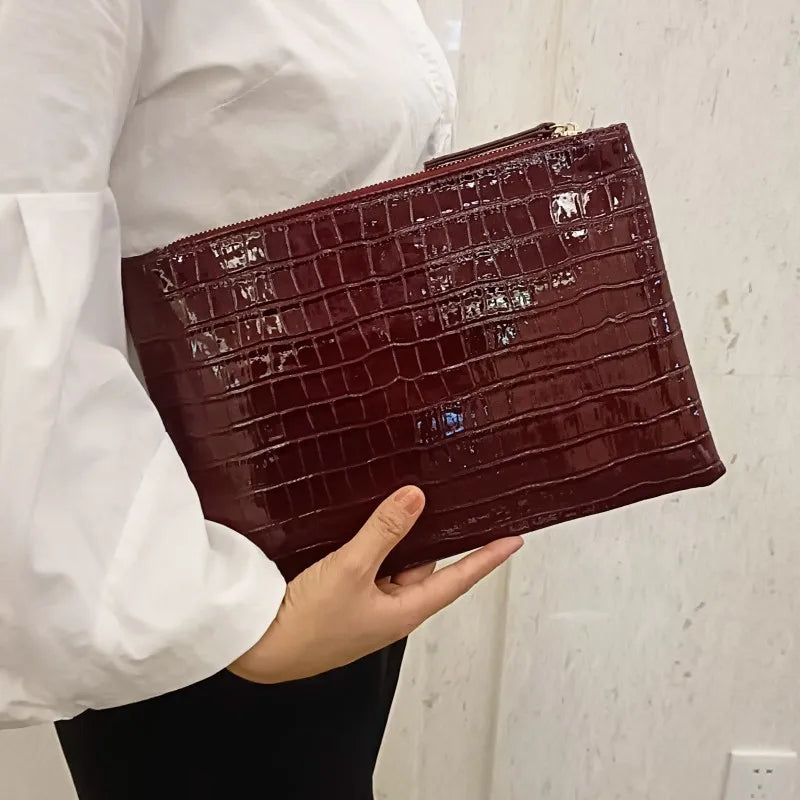 A person holds a dark burgundy alligator print clutch bag with a gold clasp.