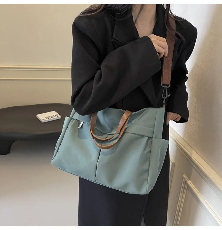 A woman holds a light blue nylon tote bag with brown leather straps against a light wall.