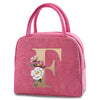A pink insulated canvas lunch bag featuring a large gold letter F and a small floral design.