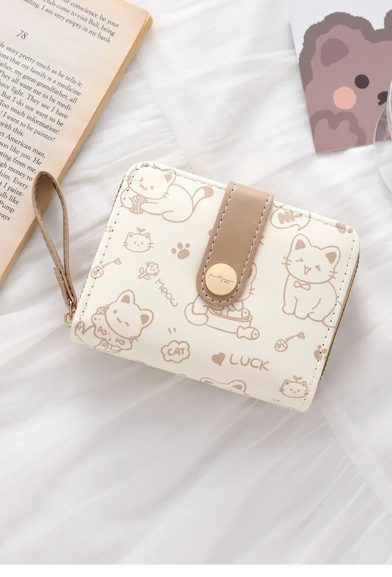 A beige cat cartoon coin purse and card holder wallet with a leather strap resting on a white fabric background.