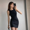 A woman wearing a black sleeveless bodycon tank dress is posing indoors.