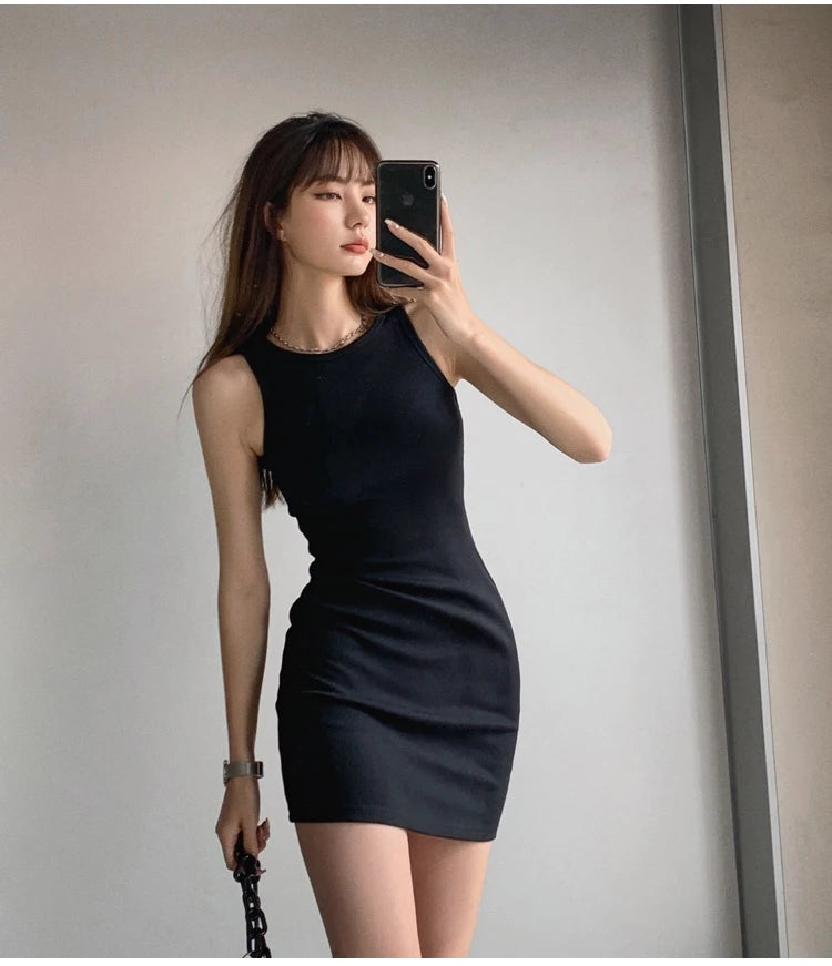 A woman wearing a black sleeveless bodycon tank dress is posing indoors.
