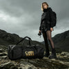 A woman wearing dark outdoor gear stands on rocks with a black storage tote labeled Carry Tools.