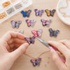 Hands are assembling colorful butterfly enamel charms for jewelry making on a wooden surface.