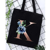 Black canvas tote bag featuring a large gold letter K with blue floral accents