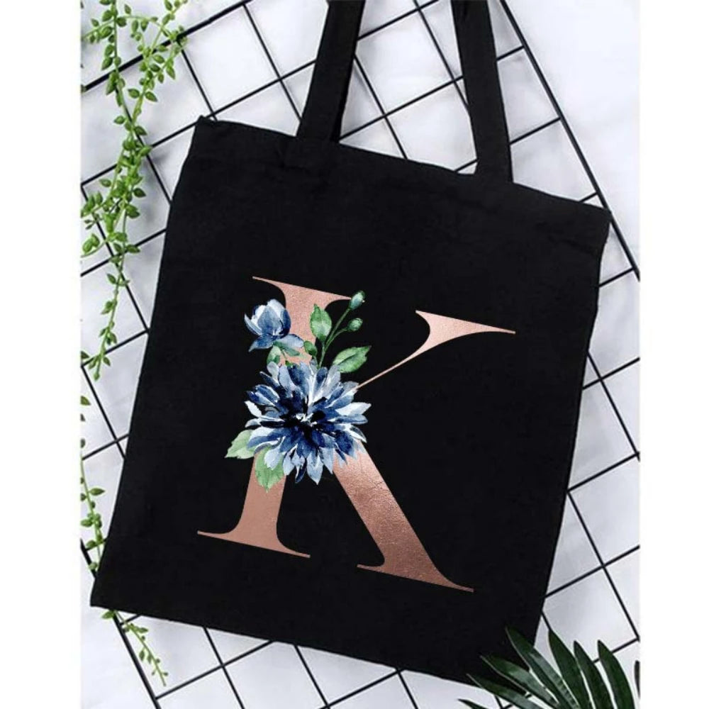 Black canvas tote bag featuring a large gold letter K with blue floral accents