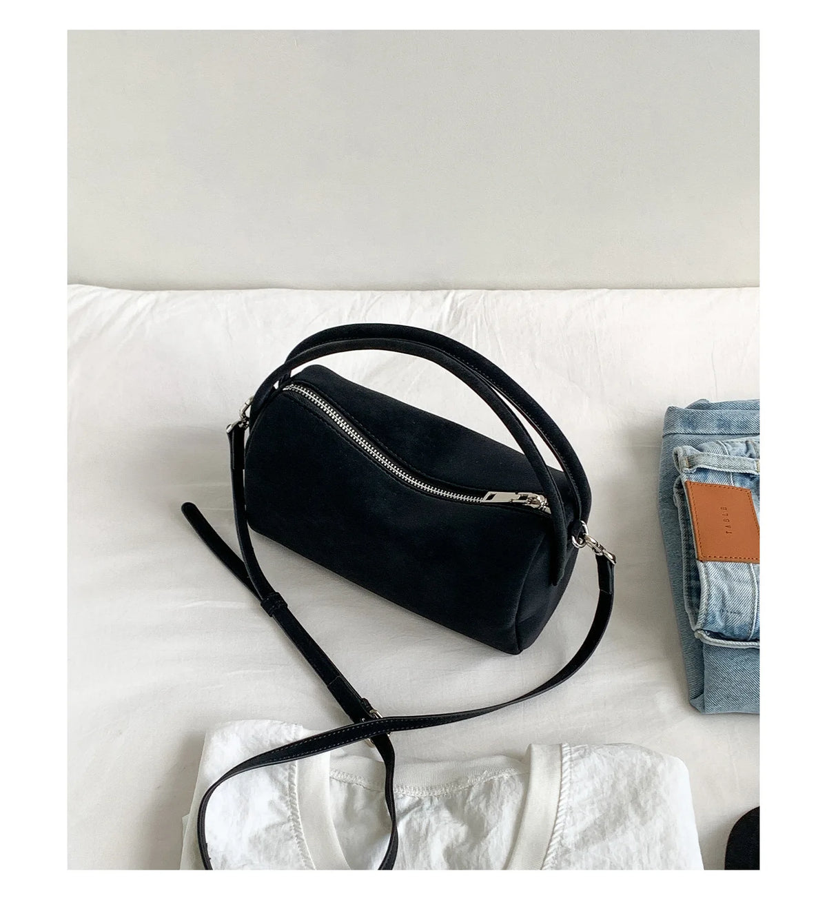 A black faux suede crossbody bag resting on a white surface next to a denim shirt.