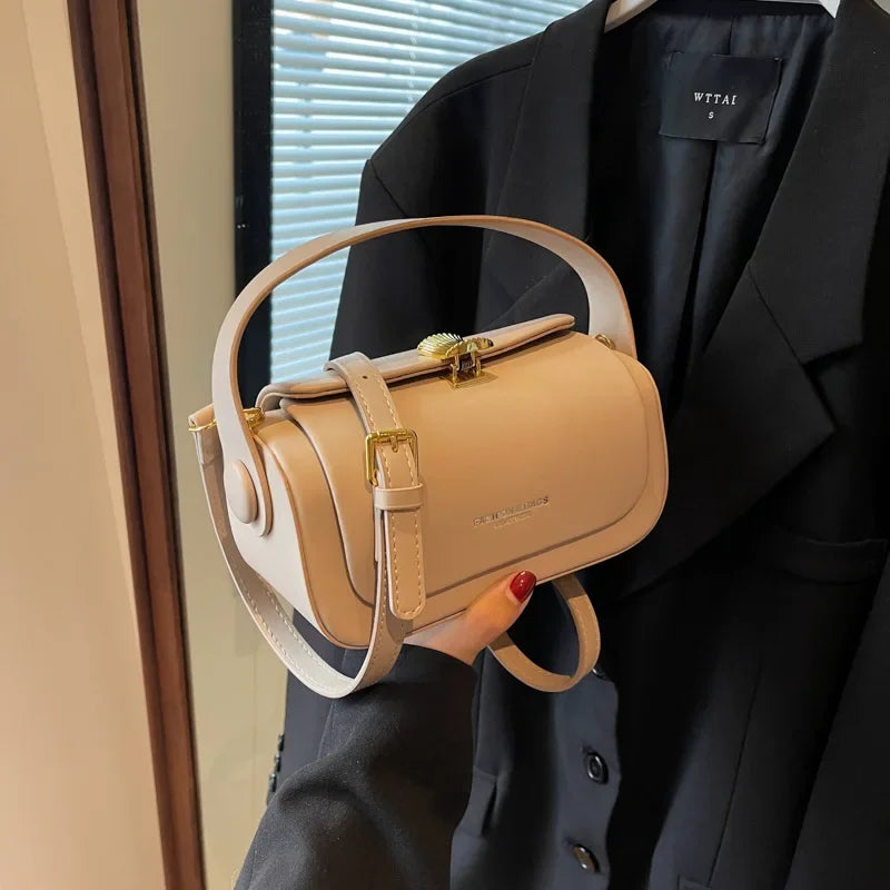 A light beige leather shoulder bag with gold hardware is shown being held against a black blazer.