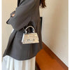A silver and gold square crossbody bag with a chain strap is shown against a gray blazer and white skirt.