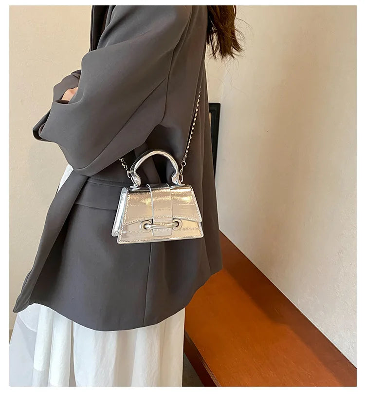 A silver and gold square crossbody bag with a chain strap is shown against a gray blazer and white skirt.