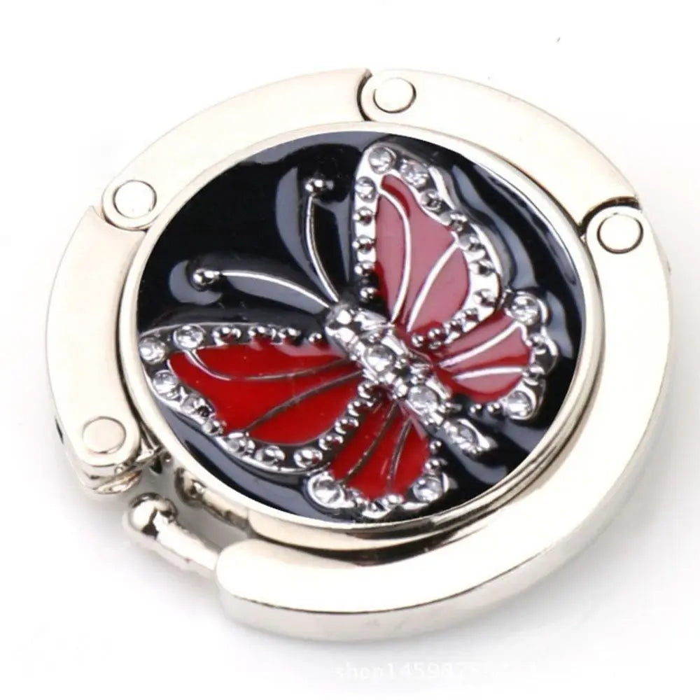 Gancho de mesa plegable de aluminio plateado con diseño de mariposa decorativa.