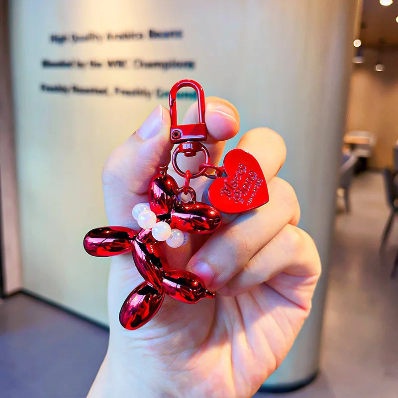 A hand holds a red balloon dog keychain charm with a silver clasp and a heart-shaped charm.