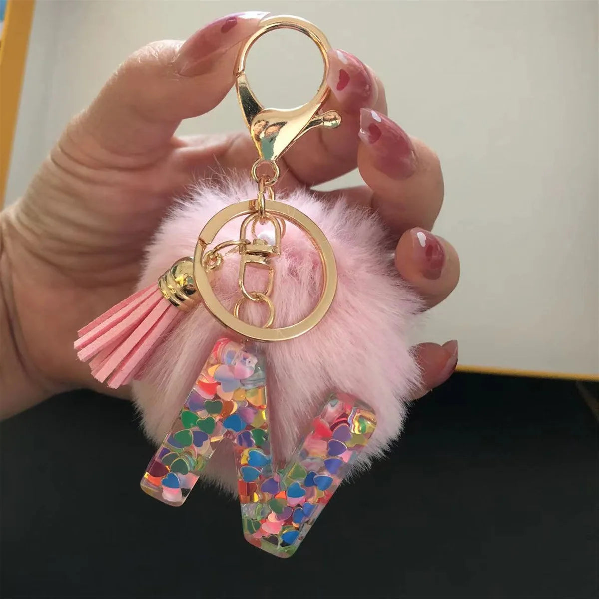 A hand holds a gold initial letter keychain featuring a shiny resin pendant and pink alphabet charm on a fluffy pink backgrou