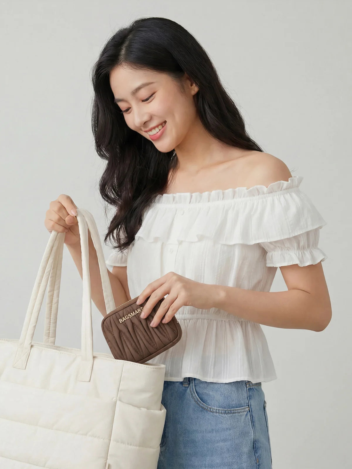 A woman holds a beige canvas tote bag and a brown coin purse against a light gray background.