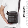 A dark brown vintage leather messenger bag with a black strap is shown being held by a person wearing a white t-shirt.