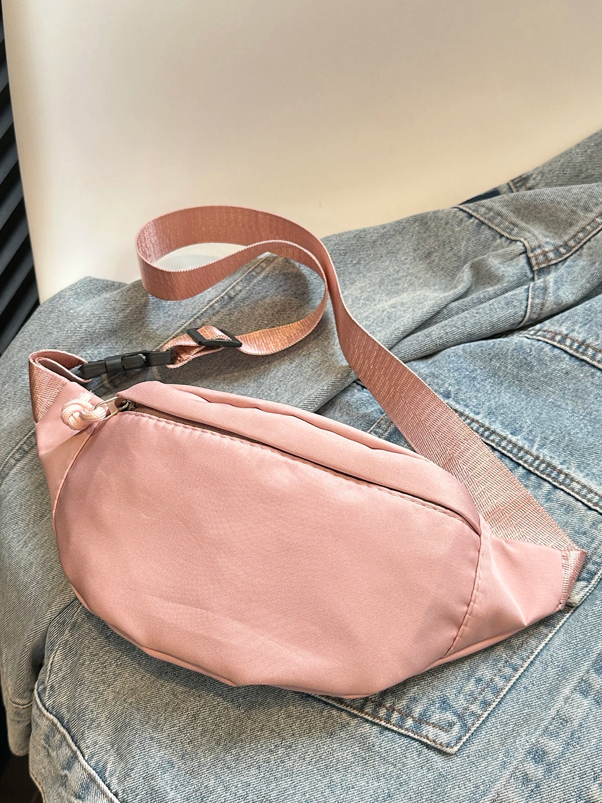 A light pink nylon crossbody sling bag with a tan strap resting on a denim jacket.