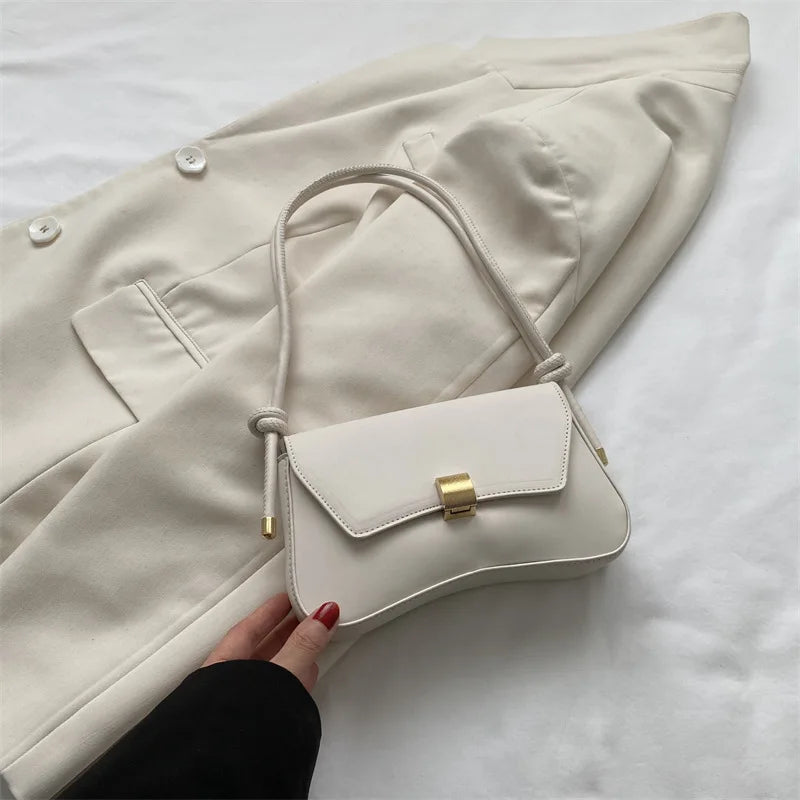 A beige leather crossbody bag with a gold clasp is shown resting on a light-colored blazer.