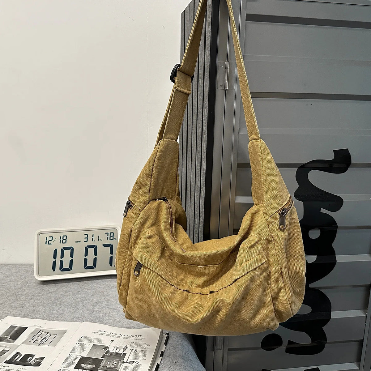 A tan cotton canvas shoulder crossbody bag hanging against a gray corrugated metal surface.