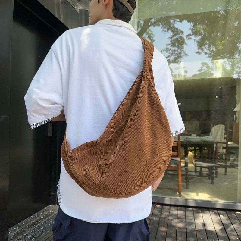 A person wearing a white shirt and dark pants is holding a brown corduroy casual crossbody shoulder bag.