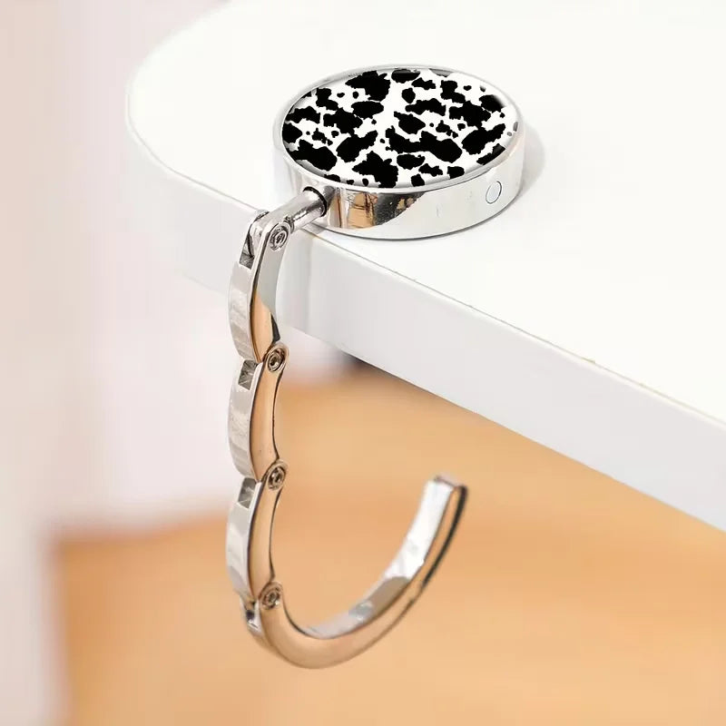 A silver zinc alloy folding handbag hook with a black and white spotted leather charm resting on a white surface.