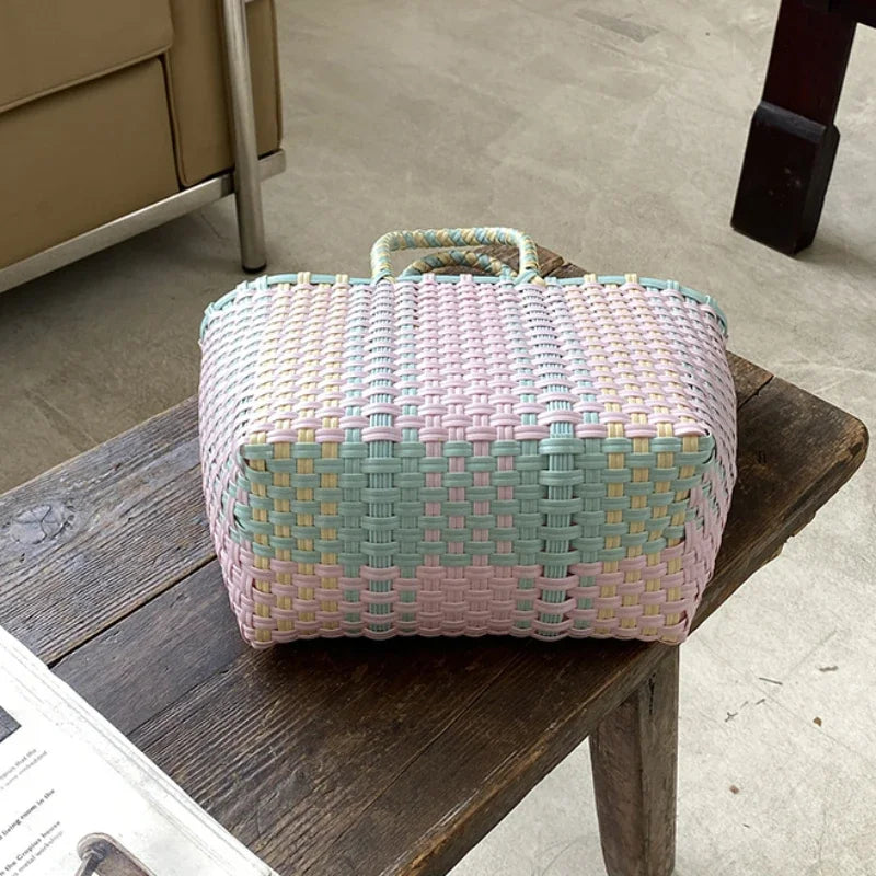 A hand woven PVC vegetable basket tote bag with pink, green, and white stripes resting on a wooden table.