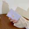 Cartera de cuero PU morado claro de tres pliegues con un dije de cinta dorada, sostenida en la mano de una persona sobre una 