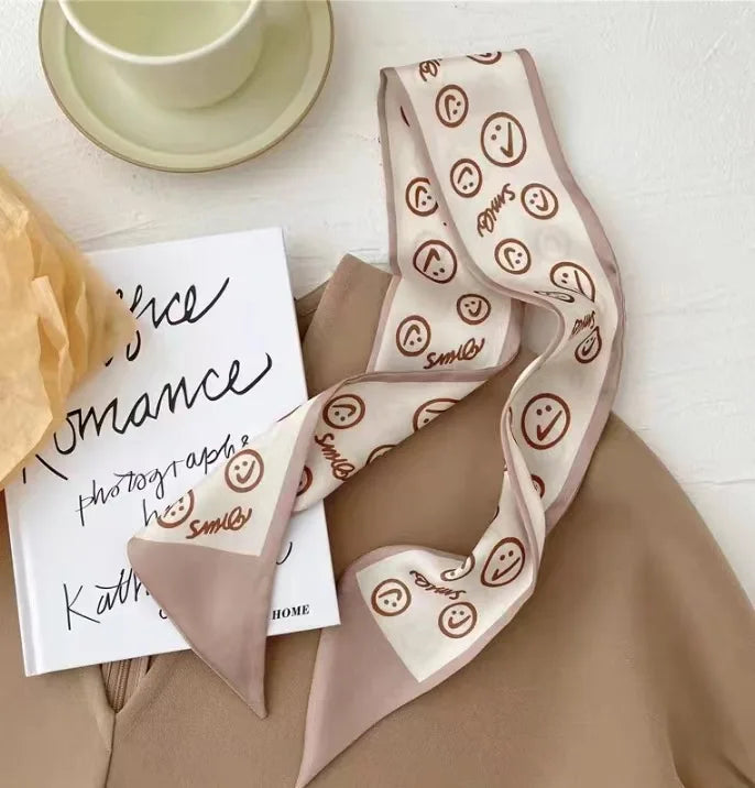 Un foulard de cheveux en soie beige avec des motifs de visages souriants, drapé à côté d'un carnet et d'une tasse à thé.