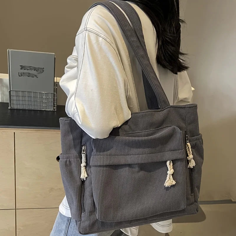 Sac cabas en velours côtelé gris avec plusieurs poches, vu de dos, porté par-dessus un t-shirt blanc.