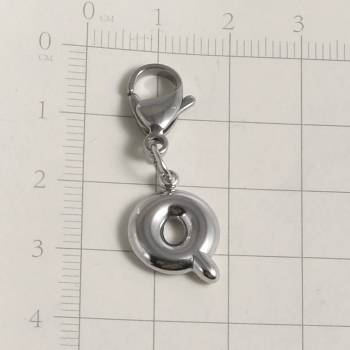 Silver stainless steel alphabet letter charm with a lobster clasp placed on a ruler grid.