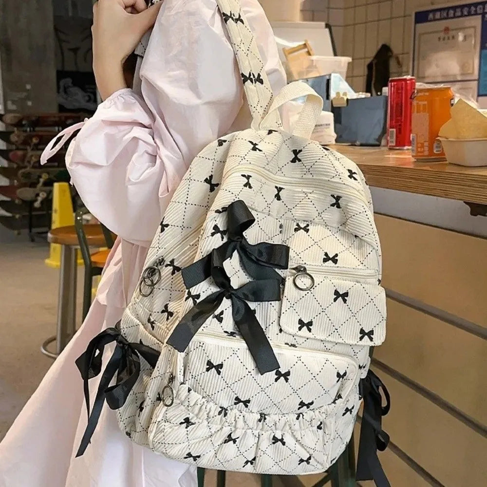 A woman holds a white backpack with black bow print details in a light pink dress.