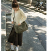 A young woman wearing a yellow knit sweater and dark skirt walks down a sidewalk carrying a large olive green canvas shoulder