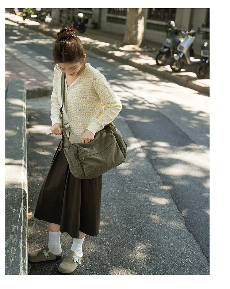 A young woman wearing a yellow knit sweater and dark skirt walks down a sidewalk carrying a large olive green canvas shoulder