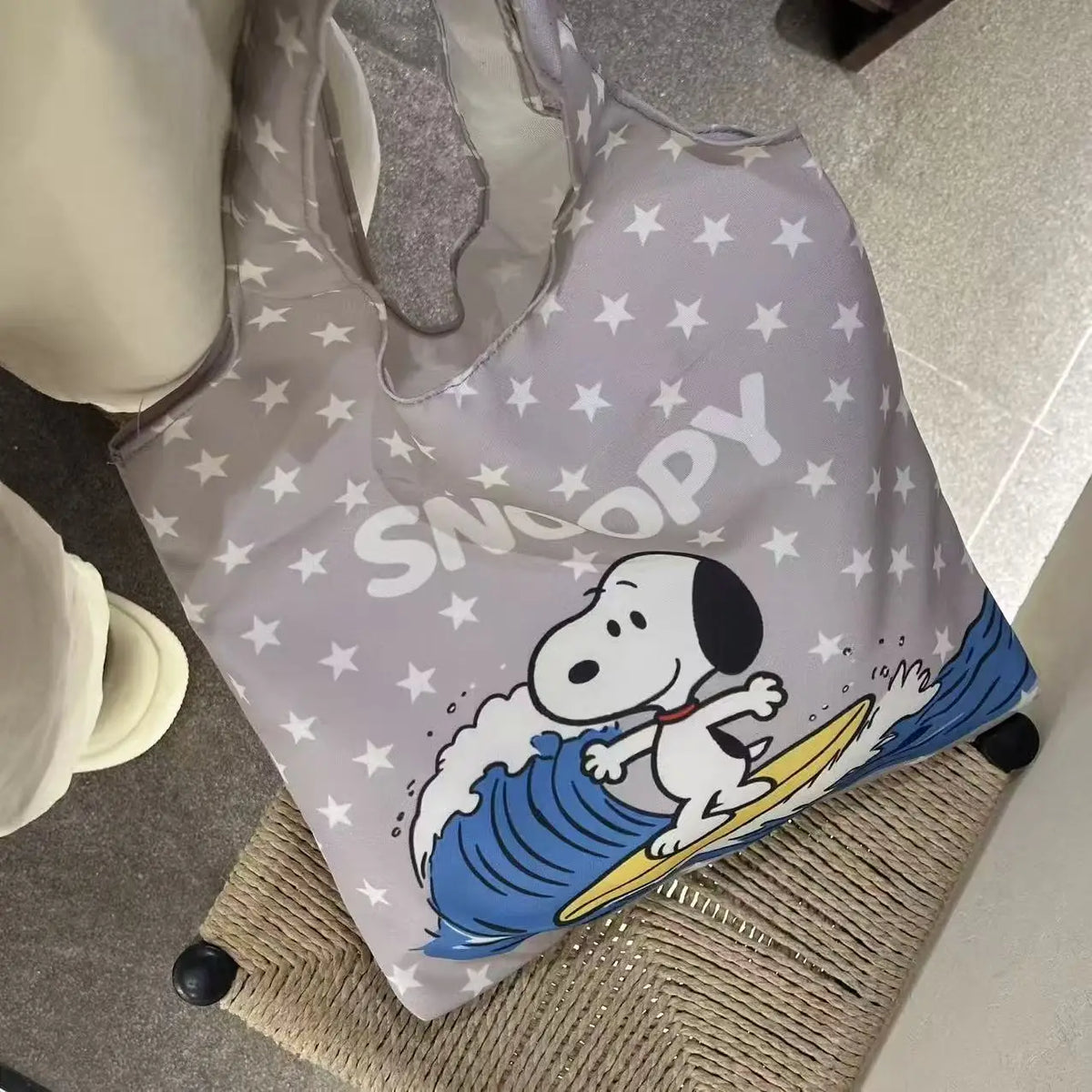 A light gray canvas shoulder bag featuring a Snoopy cartoon design with white stars and blue waves.