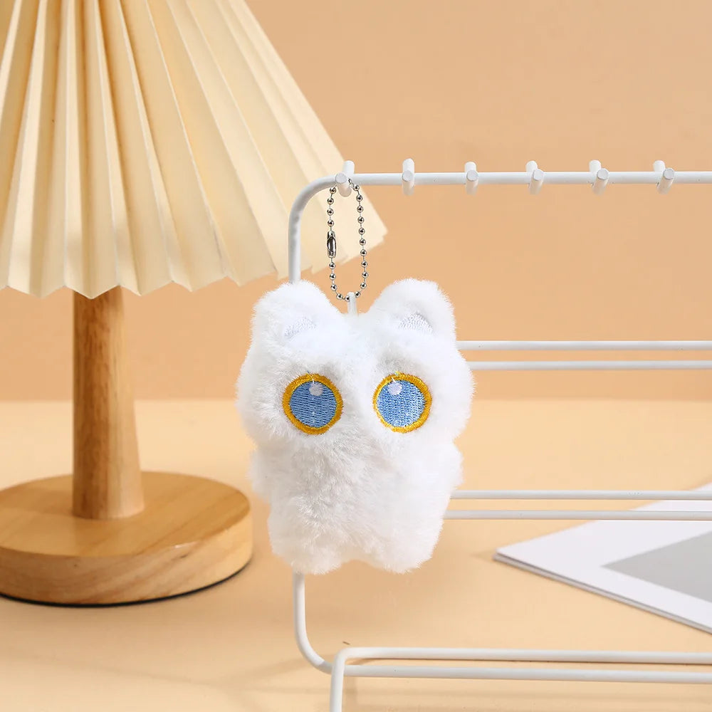 A white fluffy cat charm with blue eyes is hanging on a silver wire rack next to a wooden lamp.