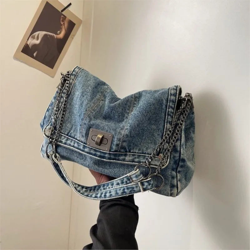 A blue denim tote bag with a silver chain strap is displayed against a white wall.