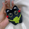 Colgante llavero de peluche de gato negro con grandes ojos amarillos y acentos verdes, sostenido en una mano contra un fondo 