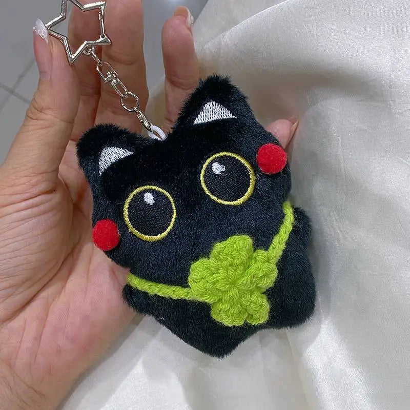 Colgante llavero de peluche de gato negro con grandes ojos amarillos y acentos verdes, sostenido en una mano contra un fondo 